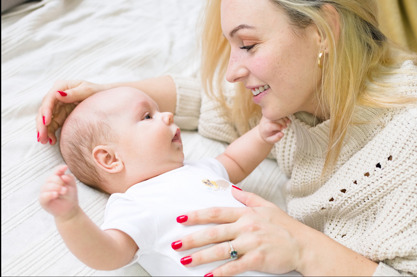 Matka z blond włosami i czerwonym manicure patrzy z uśmiechem na leżącego niemowlaka w białym body z aplikacją, na kremowym tle.