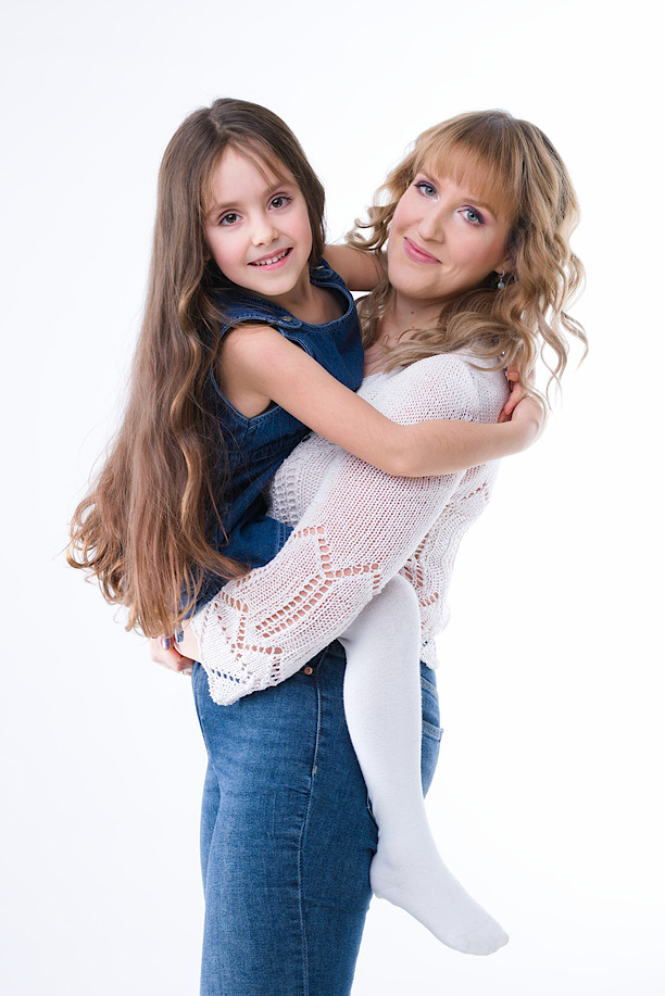 Uśmiechnięta mama w białym swetrze tuli córkę w dżinsowej sukience na białym tle. Profesjonalne oświetlenie podkreśla naturalne piękno modelek.