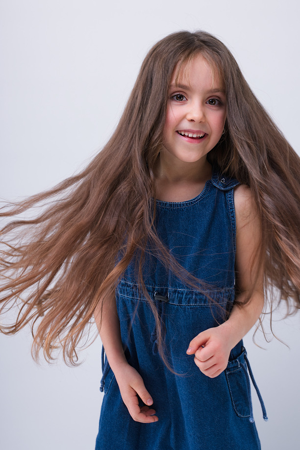 Uśmiechnięta dziewczynka z długimi brązowymi włosami w ruchu, ubrana w dżinsową sukienkę na tle jasnego studia.