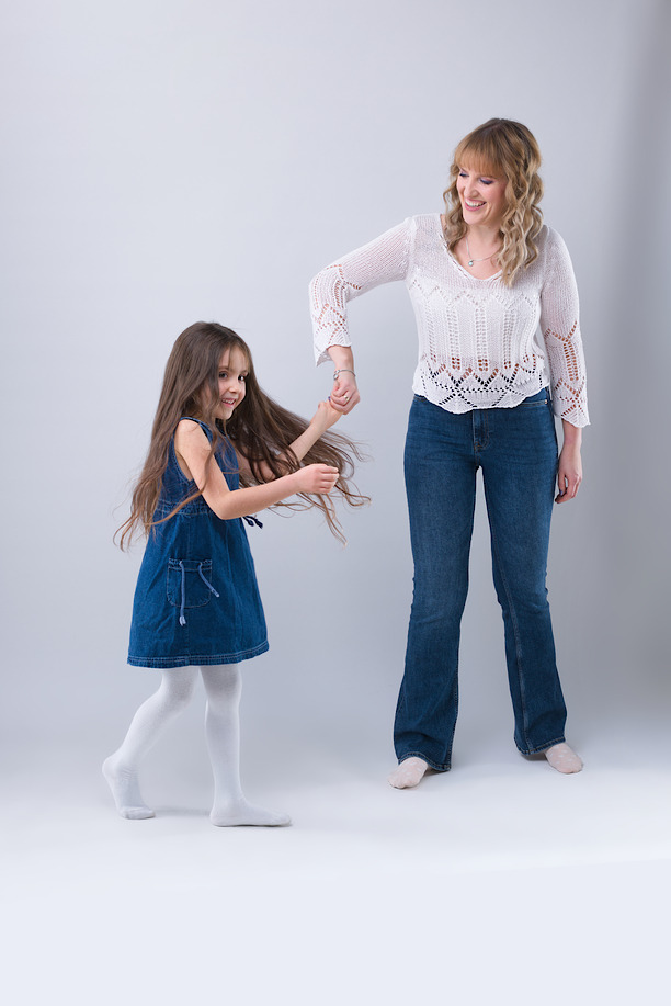 Uśmiechnięta mama w ażurowej bluzce i dżinsach prowadzi za rękę rozbawioną córeczkę w dżinsowej sukience, na jasnoszarym tle.