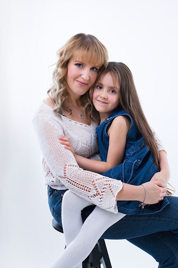 Portret matki z córką na białym tle, matka w ażurowym swetrze i jeansach, córka w dżinsowej sukience, uśmiechnięte twarze.