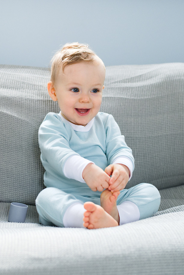 Uśmiechnięty niemowlak w błękitnym ubranku siedzi na szarym siedzisku, trzymając stópkę w dłoniach, z widocznym fragmentem szarego kubeczka obok.
