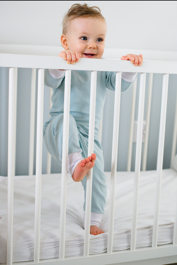 Uśmiechnięty niemowlak w błękitnym ubranku, stojący w białym łóżeczku, trzymający się szczebelków, z podniesioną jedną stopą.