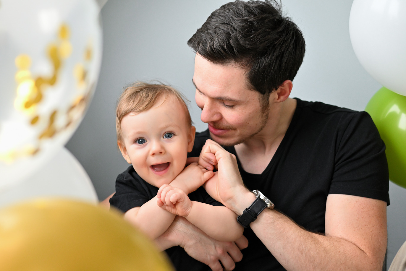 Uśmiechnięty tata w czarnej koszulce trzyma roześmianego synka na tle balonów w odcieniach bieli, złota i zieleni.