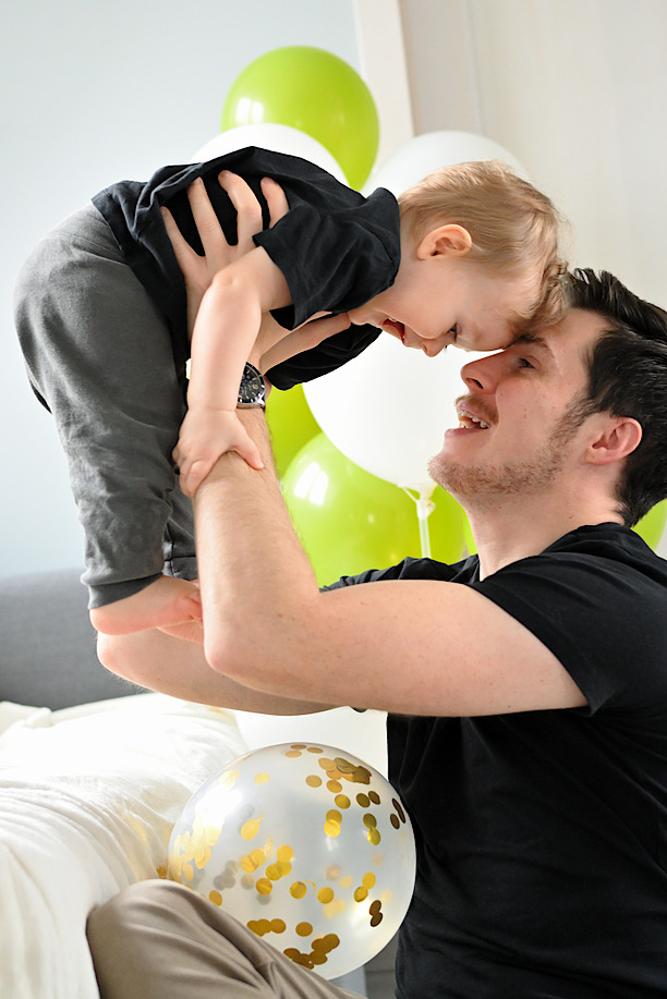 Uśmiechnięty ojciec podnosi w górę roześmianego synka, w tle zielone i białe balony, jeden przezroczysty z konfetti leży na łóżku.