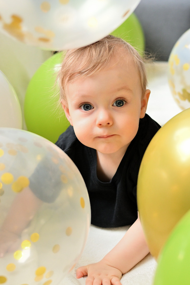 Niemowlę w czarnym ubranku leżące na białym tle, otoczone przezroczystymi, zielonymi i złotymi balonami z konfetti, patrzy w obiektyw.