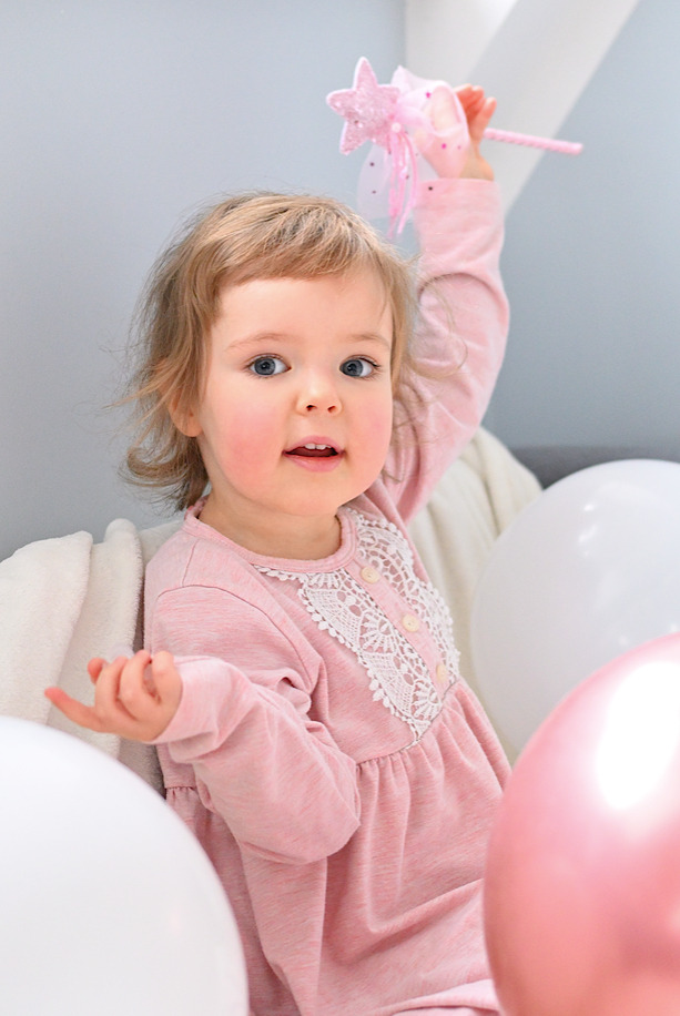 Uśmiechnięta dziewczynka w różowej sukience z koronką, trzymająca różdżkę z gwiazdką, otoczona białymi i różowymi balonami.