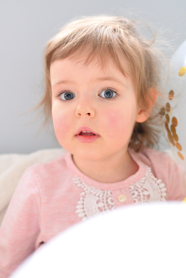 Portret uroczej, jasnowłosej dziewczynki z niebieskimi oczami, ubrana w różową bluzkę z koronkowym wykończeniem, częściowo zasłonięta białym balonem z złotymi konfetti.