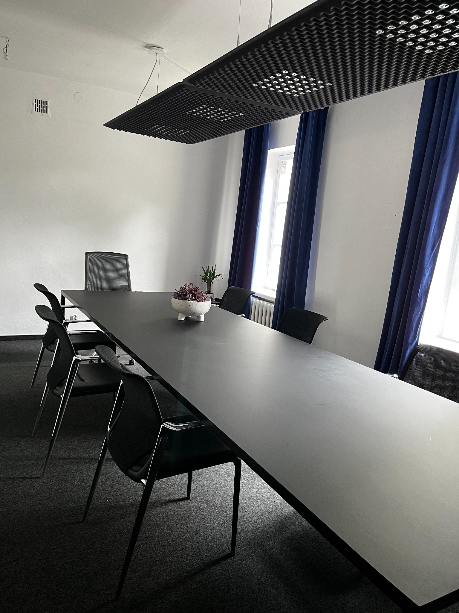 Długa, czarna sala konferencyjna z krzesłami, białymi ścianami, niebieskimi zasłonami i modernistyczną lampą sufitową.