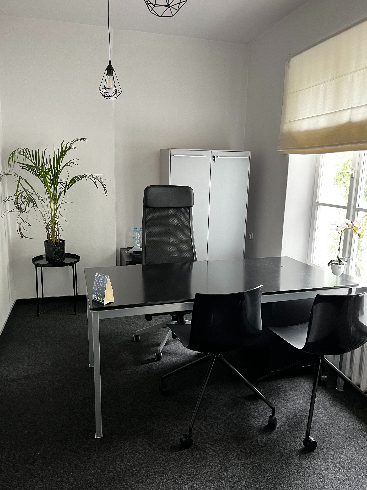 Nowoczesne, minimalistyczne biuro z czarnym biurkiem, krzesłami, rośliną doniczkową na stoliku i białą szafą w tle, oświetlone naturalnym światłem z okna z roletą rzymską.