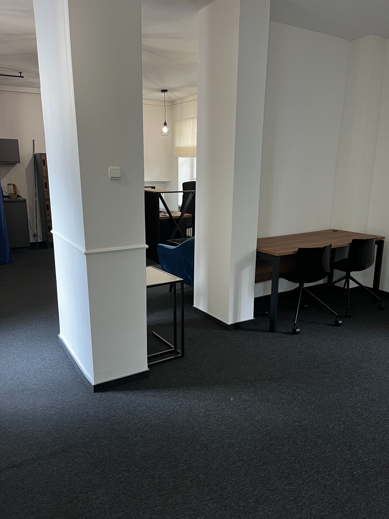 Nowoczesna aranżacja przestrzeni biurowej z biurkami, krzesłami na kółkach, minimalistycznym oświetleniem i ciemną wykładziną podłogową, widziana przez białe filary architektoniczne.