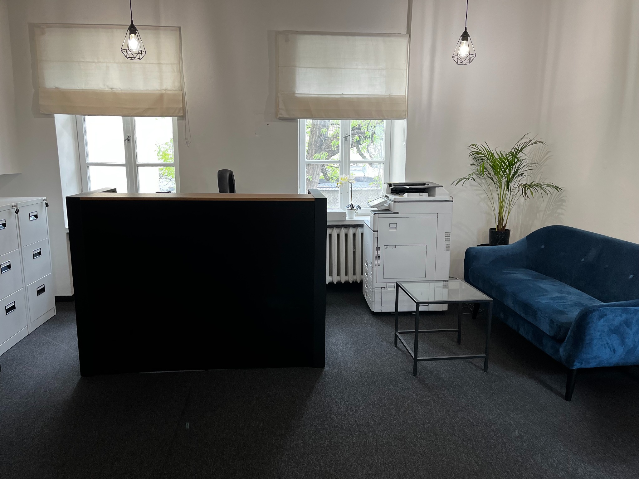 Nowoczesne wnętrze biurowe z recepcją, drukarką, sofą i rośliną doniczkową, oświetlone wiszącymi lampami z geometrycznymi kloszami.