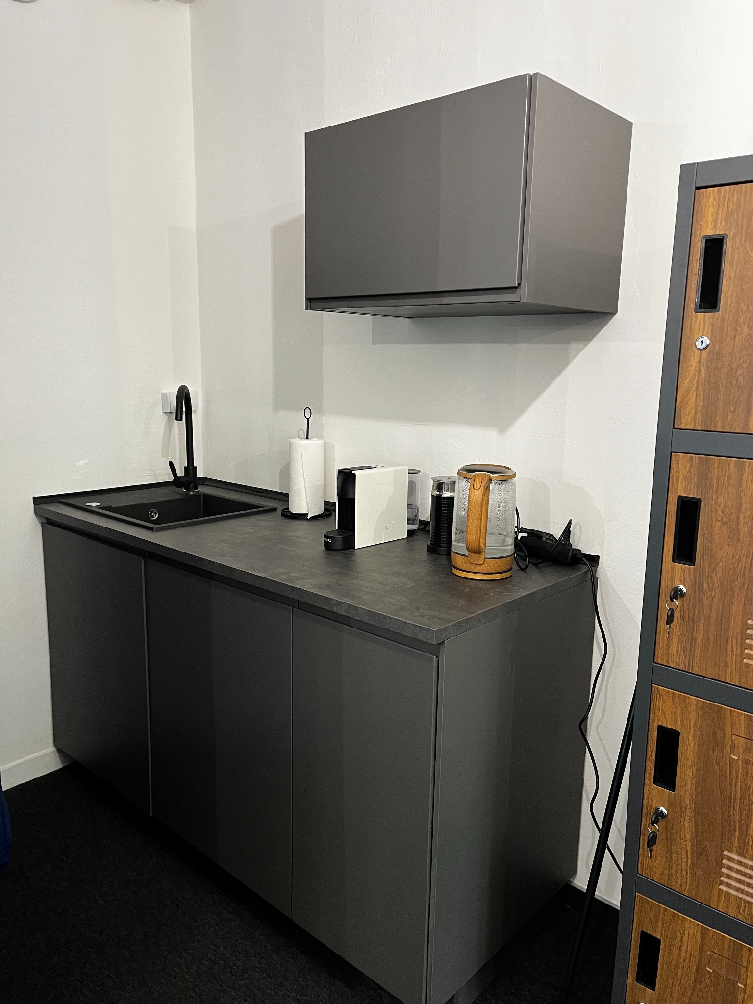 Kącik kuchenny w biurze: szare szafki pod zlewem z czarną armaturą, ekspres do kawy, czajnik elektryczny i szafka wisząca w tym samym odcieniu szarości, obok szafki pracownicze zamykane na klucz.