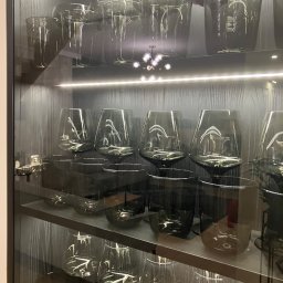 Perfect - firma sprzątająca Gdańsk Sopot Gdynia - Szklane naczynia, w tym kieliszki i szklanki, prezentowane w podświetlanej, czarnej witrynie w Gdyni. Naczynia są czyste, ułożone na trzech półkach.