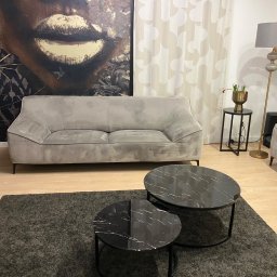 Perfect - firma sprzątająca Gdańsk Sopot Gdynia - Szara, dwuosobowa sofa na tle tapety z motywem twarzy, uzupełniona o dwa okrągłe stoliki kawowe z czarnymi blatami imitującymi marmur i szary dywan.