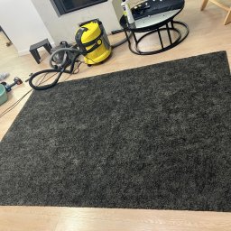 Perfect - firma sprzątająca Gdańsk Sopot Gdynia - Ciemnoszary dywan z widocznym odkurzaczem piorącym Karcher w trakcie czyszczenia na jasnej drewnianej podłodze, z elementami wyposażenia wnętrza w tle.