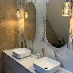 Perfect - firma sprzątająca Gdańsk Sopot Gdynia - Nowoczesna łazienka z dwoma umywalkami nablatowymi, złotymi bateriami i owalnymi lustrami podświetlonymi dekoracyjnymi lampami wiszącymi. Ściany wyłożone płytkami imitującymi marmur i drewno...