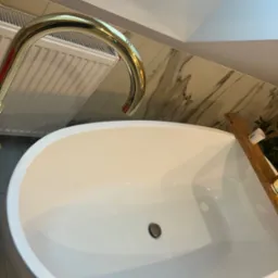 Owalna, biała wanna z złotą baterią, marmurowe płytki na ścianie, drewniana półka z akcesoriami do kąpieli, widok z góry.