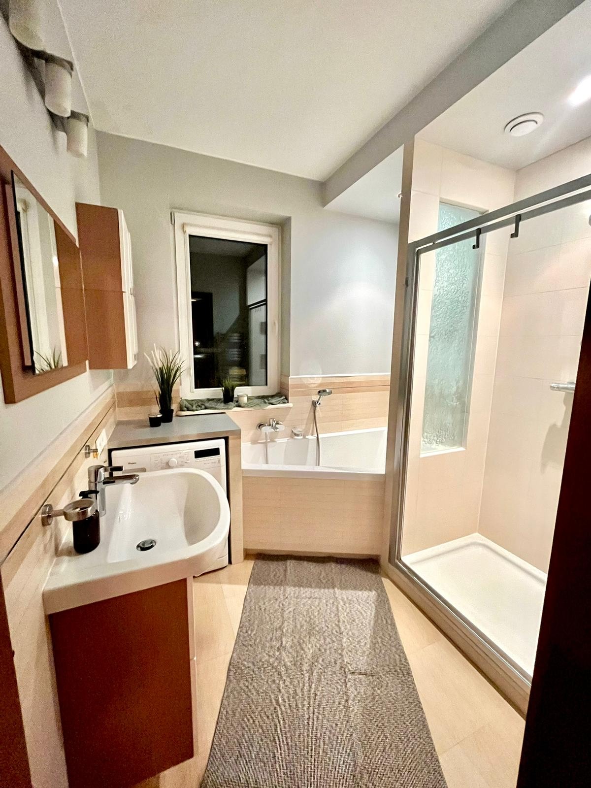 Nowoczesna łazienka z pralką pod blatem, wanną i kabiną prysznicową, utrzymana w jasnej kolorystyce z akcentami brązu i szarości, oświetlona ciepłym światłem.