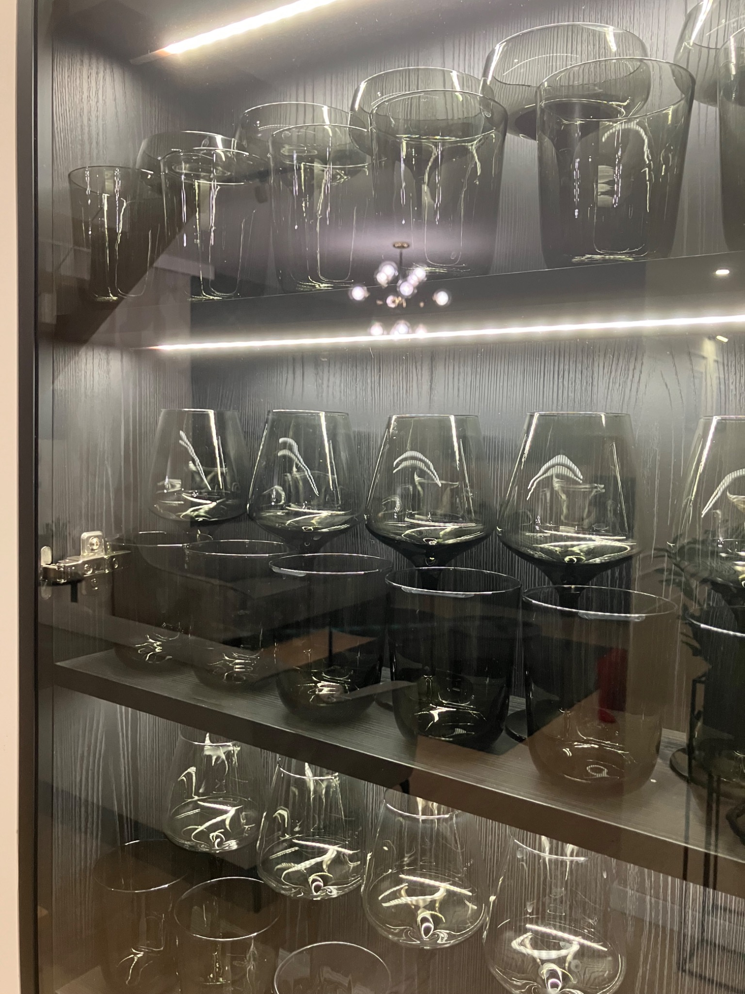Szklane naczynia, w tym kieliszki i szklanki, prezentowane w podświetlanej, czarnej witrynie w Gdyni. Naczynia są czyste, ułożone na trzech półkach.