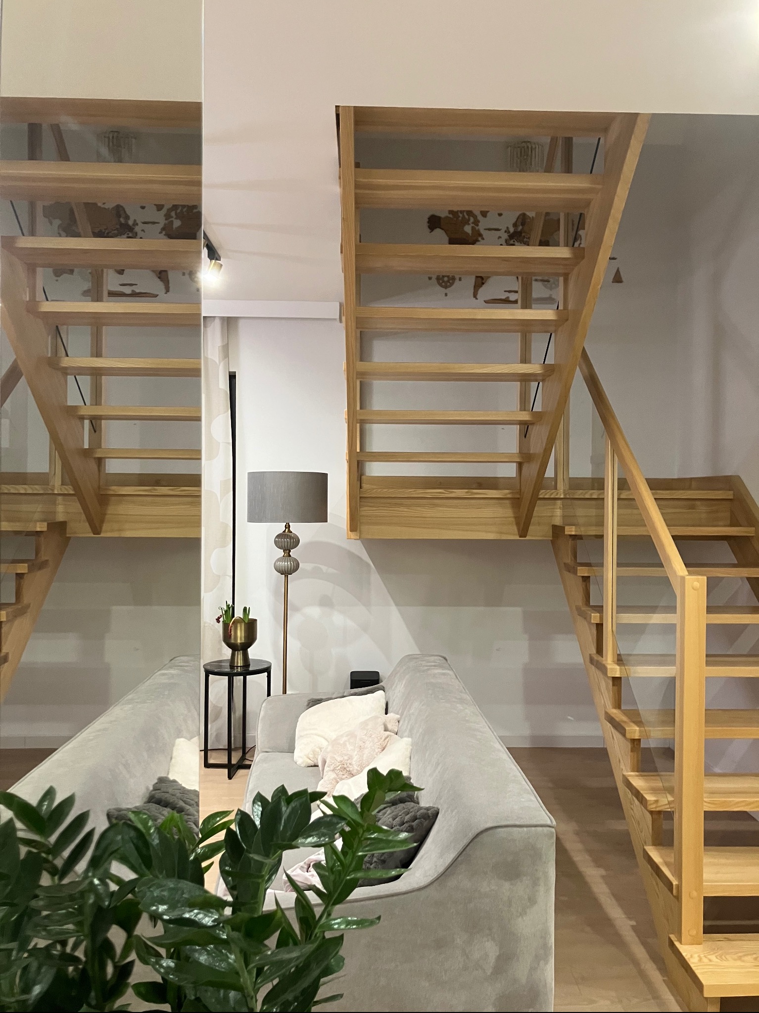 Jasne, drewniane schody w nowoczesnym wnętrzu, odbijające się w lustrze, z szarą sofą i zieloną rośliną na pierwszym planie.