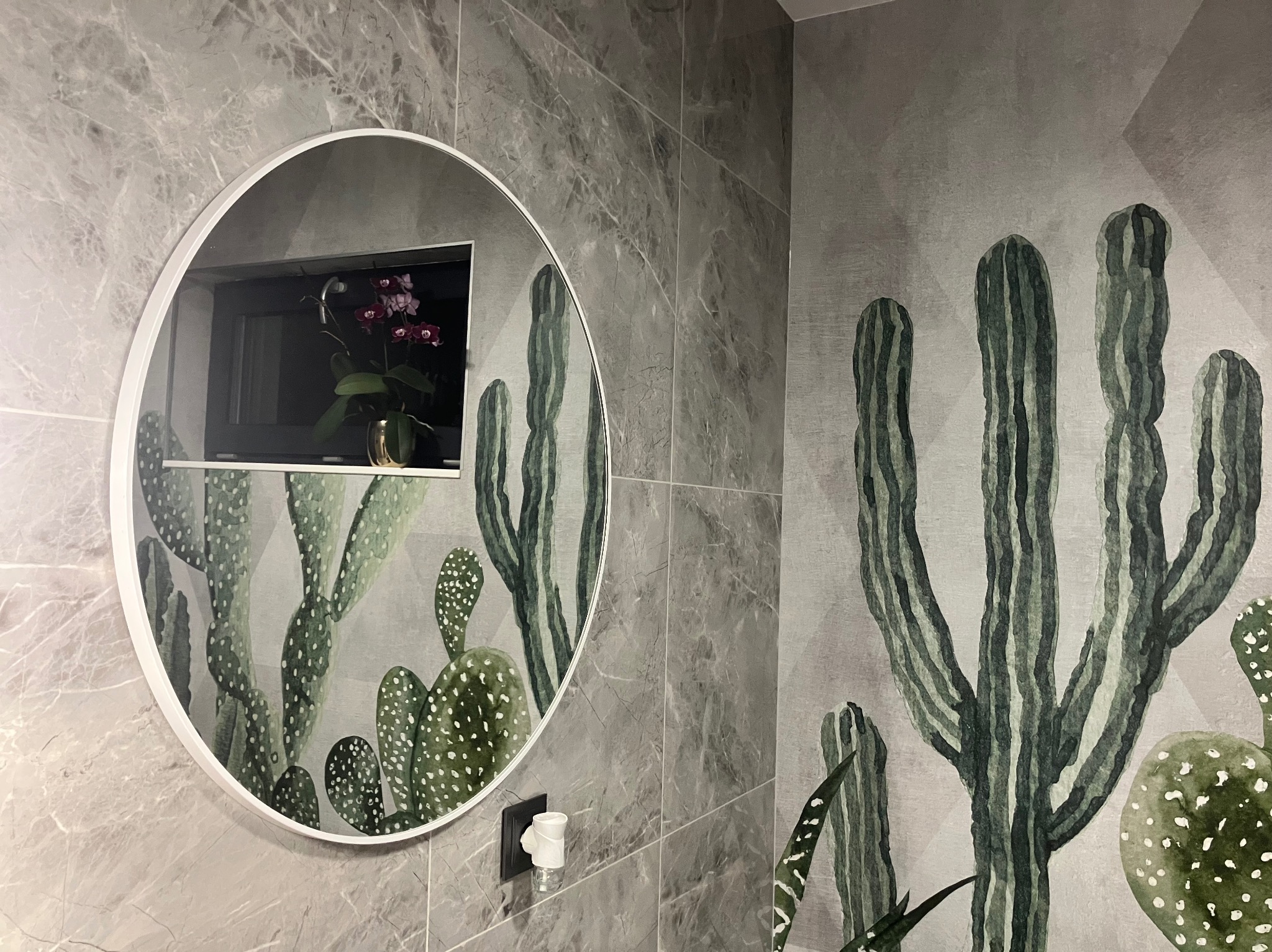 Wnętrze łazienki z szarą marmurową płytką, okrągłym lustrem odbijającym kwiaty w oknie i tapetą z motywem kaktusów.