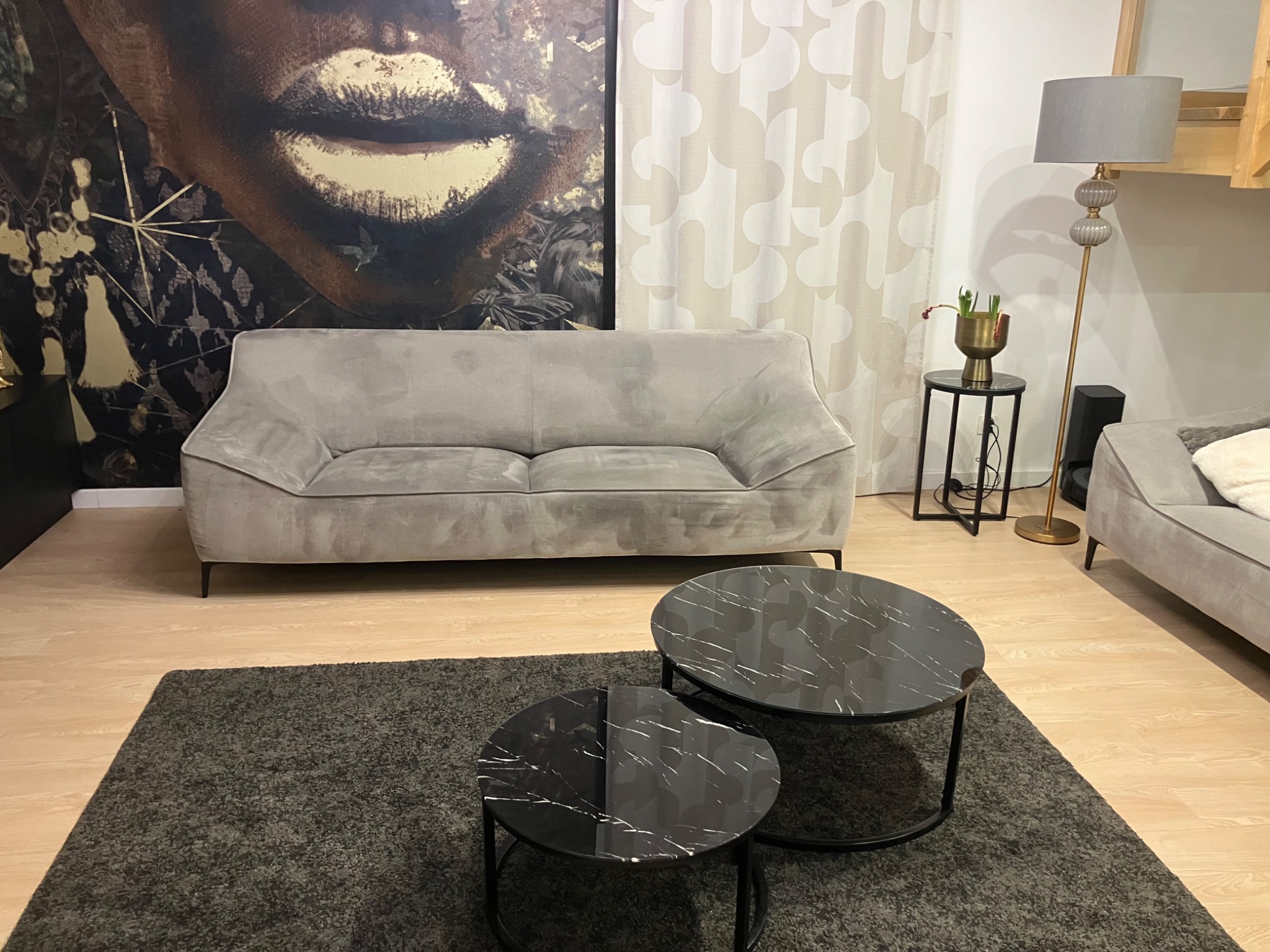 Szara, dwuosobowa sofa na tle tapety z motywem twarzy, uzupełniona o dwa okrągłe stoliki kawowe z czarnymi blatami imitującymi marmur i szary dywan.