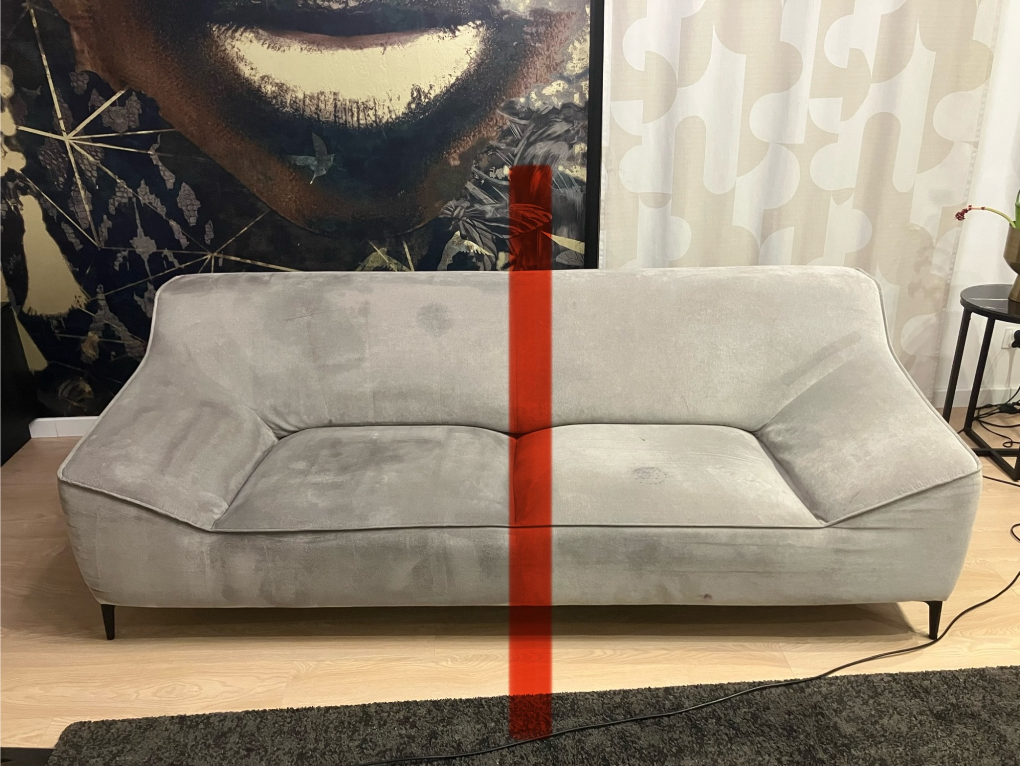 Dwuosobowa szara sofa z widocznymi plamami, podzielona w pionie czerwoną linią, na tle wzorzystej tapety i zasłon.