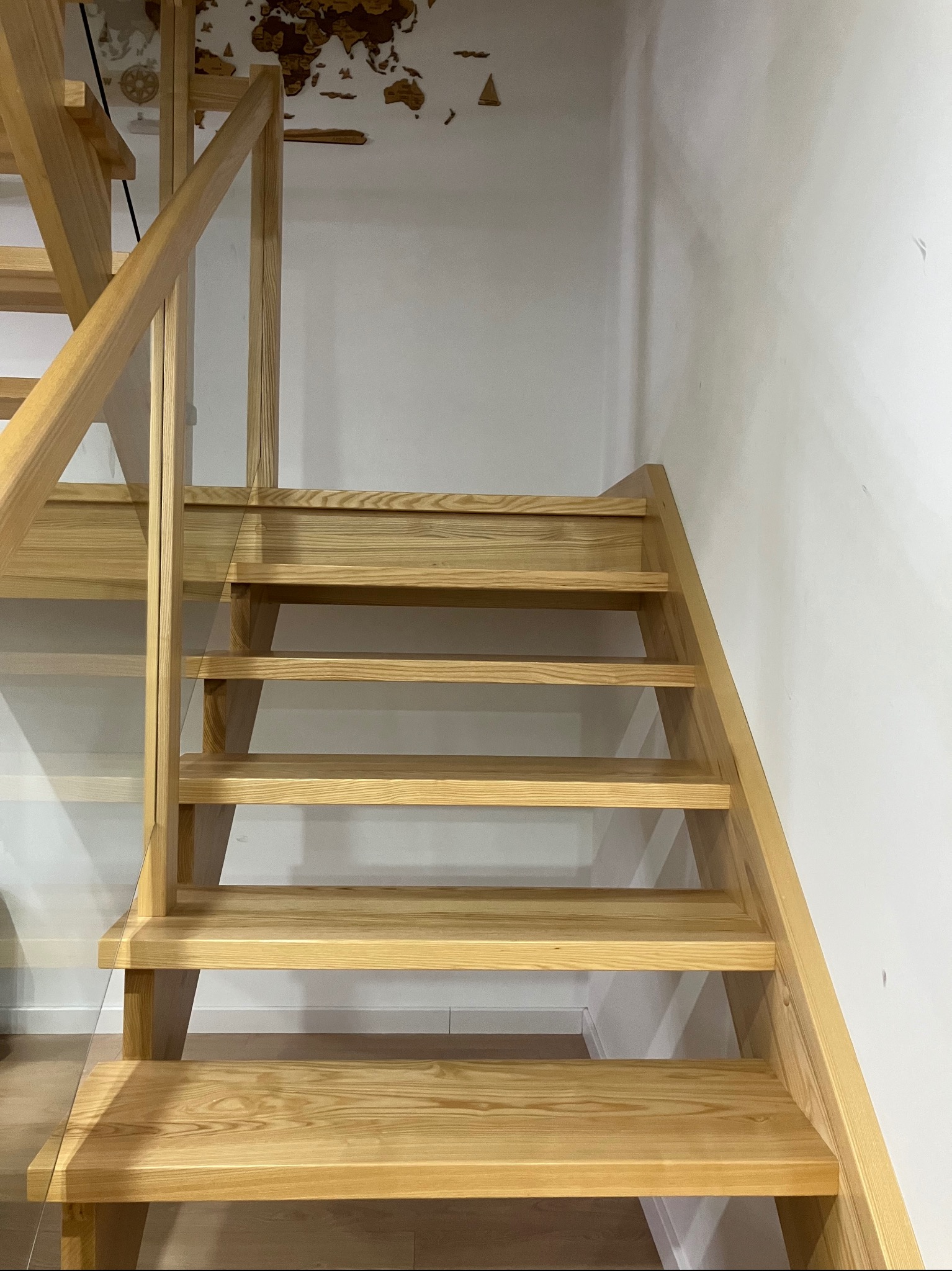 Jasne, drewniane schody z przeszkloną barierką boczną, prowadzące w górę, na białym tle ściany z dekoracją mapy świata.