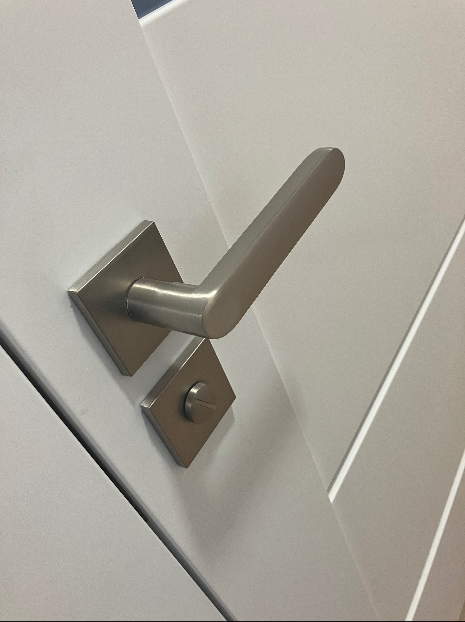 Nowoczesne, szczotkowane, metalowe okucie drzwiowe na białych, panelowych drzwiach wewnętrznych z subtelnym, geometrycznym wzorem.