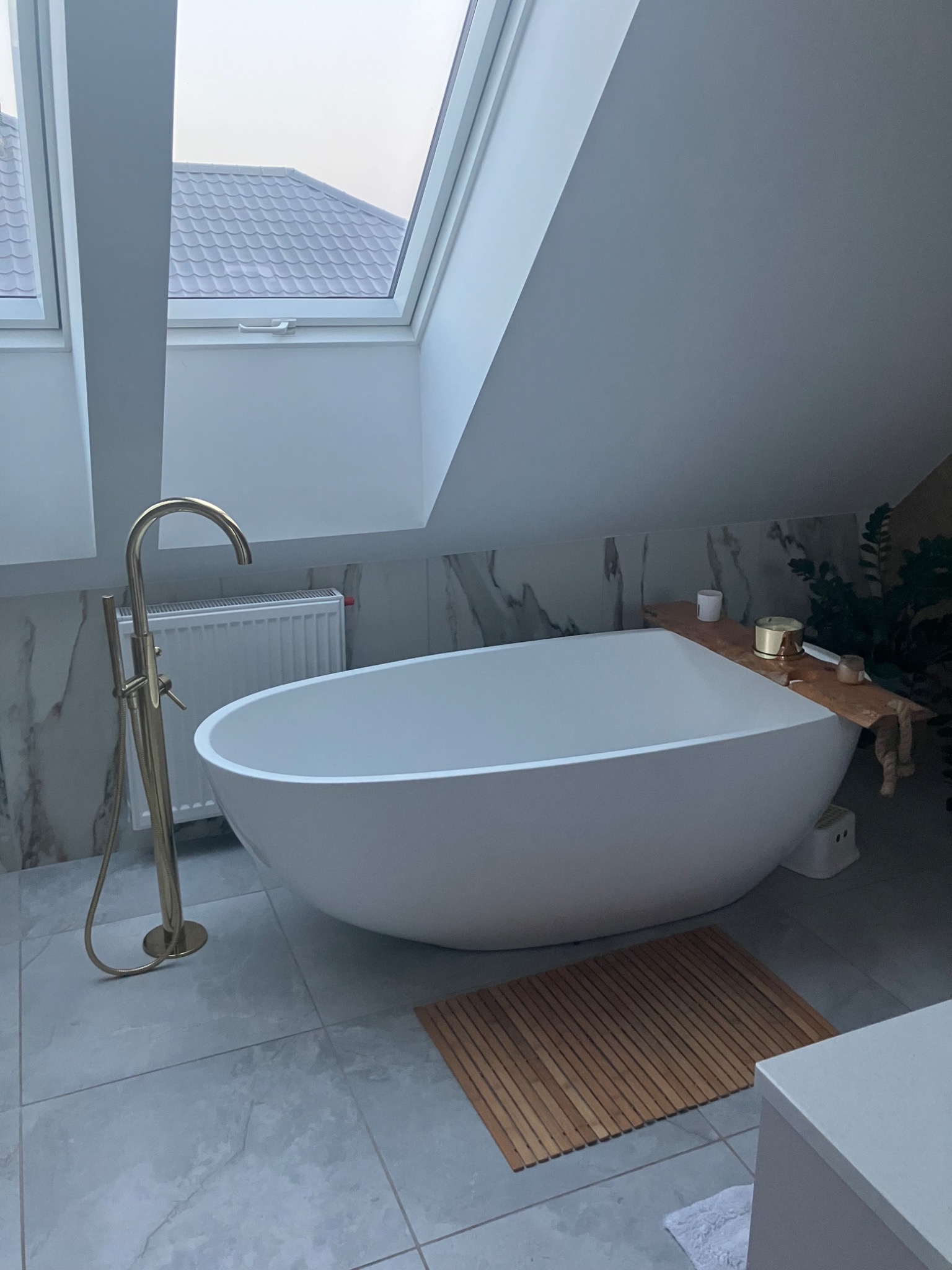 Nowoczesna, wolnostojąca wanna w jasnej łazience na poddaszu z oknem dachowym, złota bateria podłogowa, drewniana mata podłogowa.