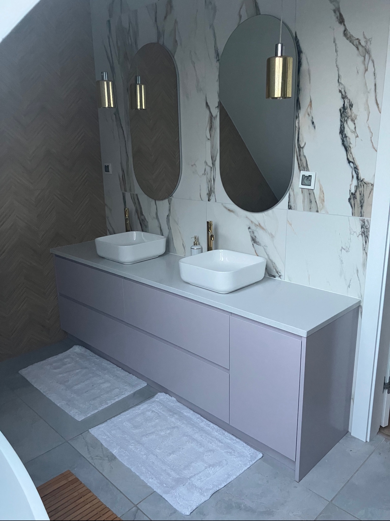 Nowoczesna łazienka z podwójną umywalką nablatową, podświetlanymi lustrami owalnymi, marmurowymi płytkami ściennymi i drewnianą mozaiką w jodełkę.