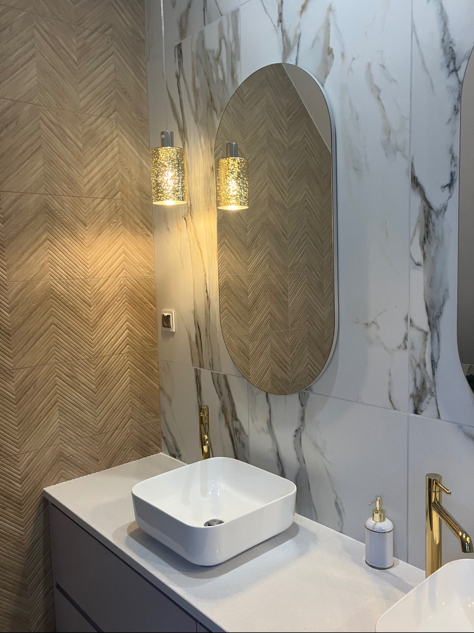 Nowoczesna łazienka z białą, kwadratową umywalką, złotą armaturą i lustrem w kształcie zaokrąglonego prostokąta, oświetlona wiszącymi lampami z efektem brokatu, na tle płytek imitujących drewno...