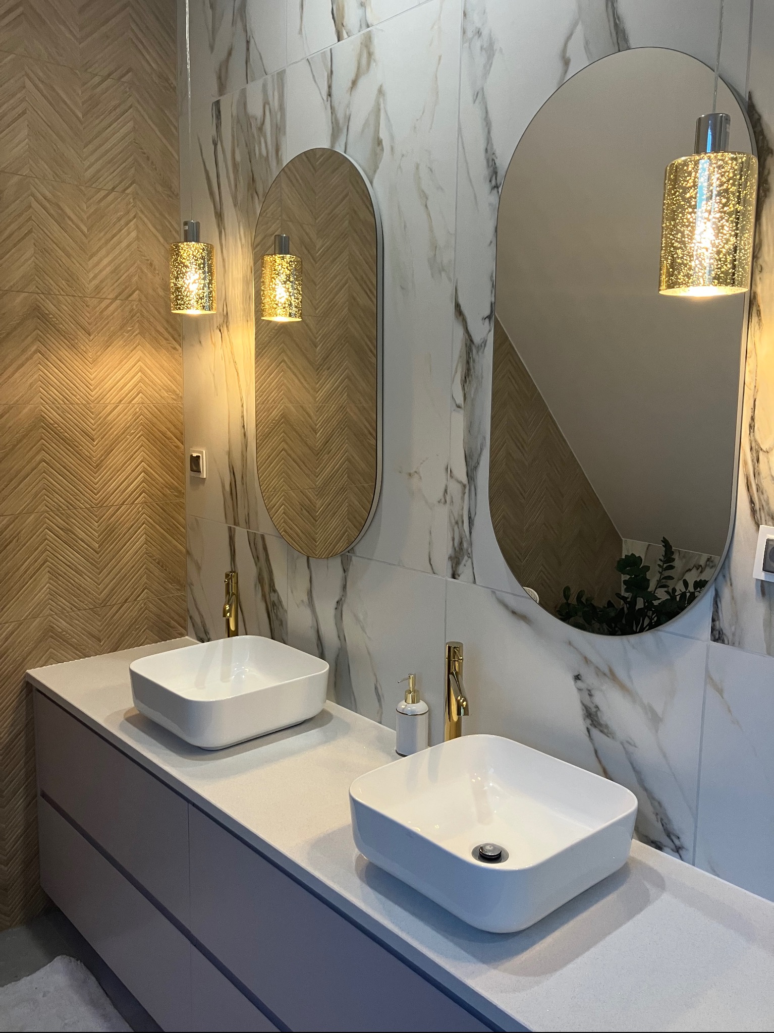 Nowoczesna łazienka z dwoma umywalkami nablatowymi, złotymi bateriami i owalnymi lustrami podświetlonymi dekoracyjnymi lampami wiszącymi. Ściany wyłożone płytkami imitującymi marmur i drewno...