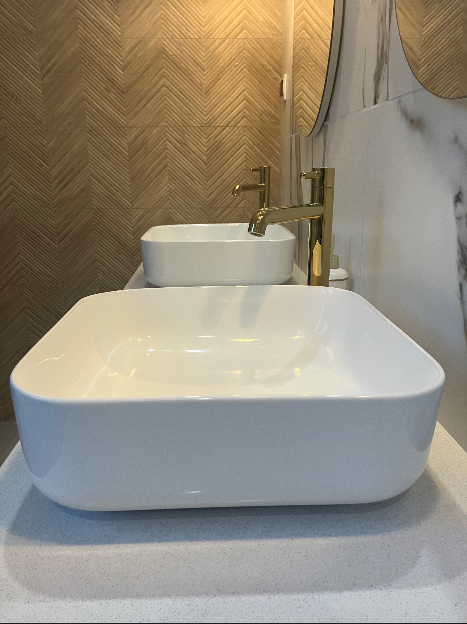 Dwie białe, kwadratowe umywalki nablatowe z bateriami w kolorze złotym, na tle drewnianej ściany ułożonej w jodełkę i fragmentu marmurowej ściany z okrągłym lustrem.