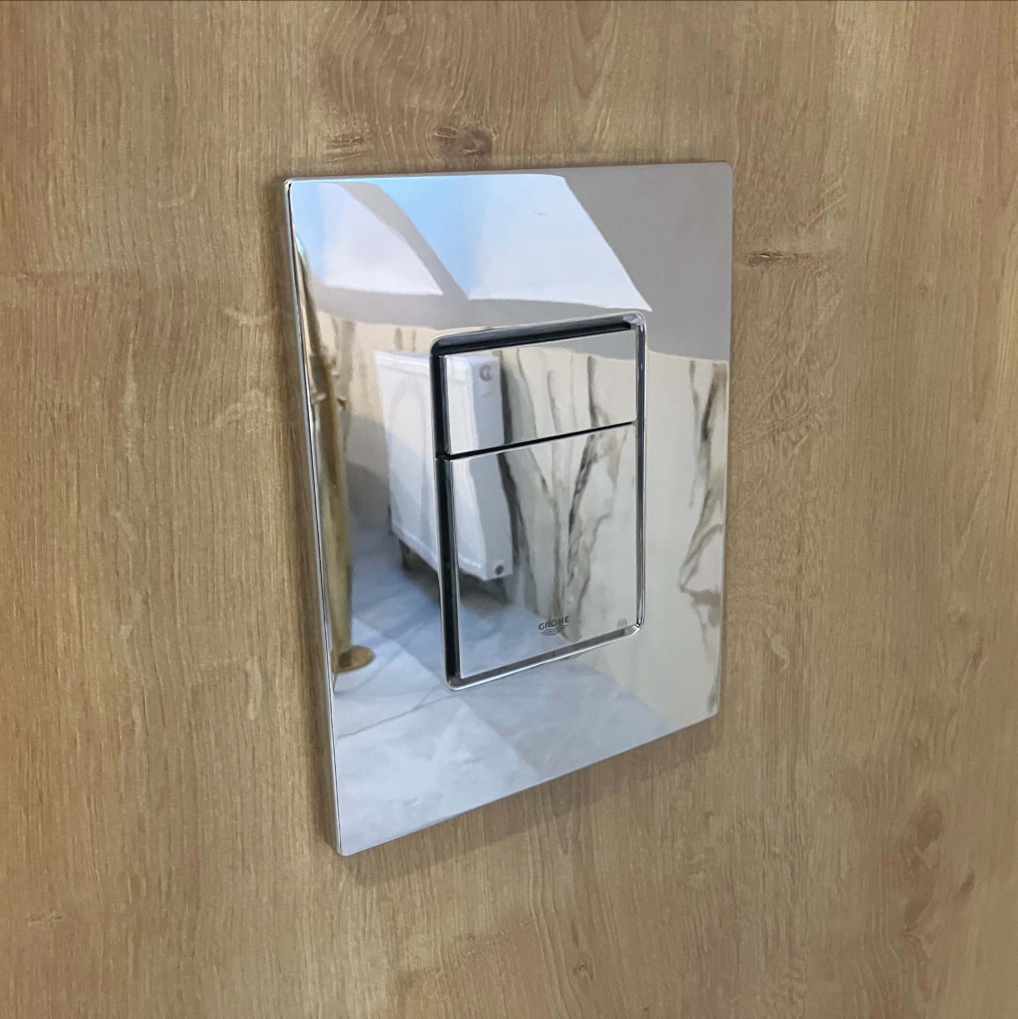 Błyszczący, chromowany przycisk spłuczki toalety marki Grohe zamontowany na drewnianej ścianie. W odbiciu widać fragment toalety i podłogi.