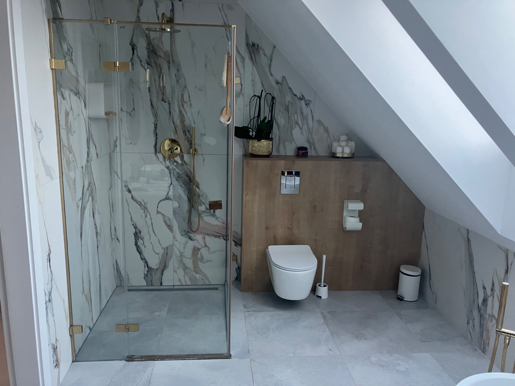 Wykończona łazienka na poddaszu z kabiną prysznicową ze złotymi okuciami i marmurowymi płytkami, wiszącą toaletą oraz drewnianą zabudową.