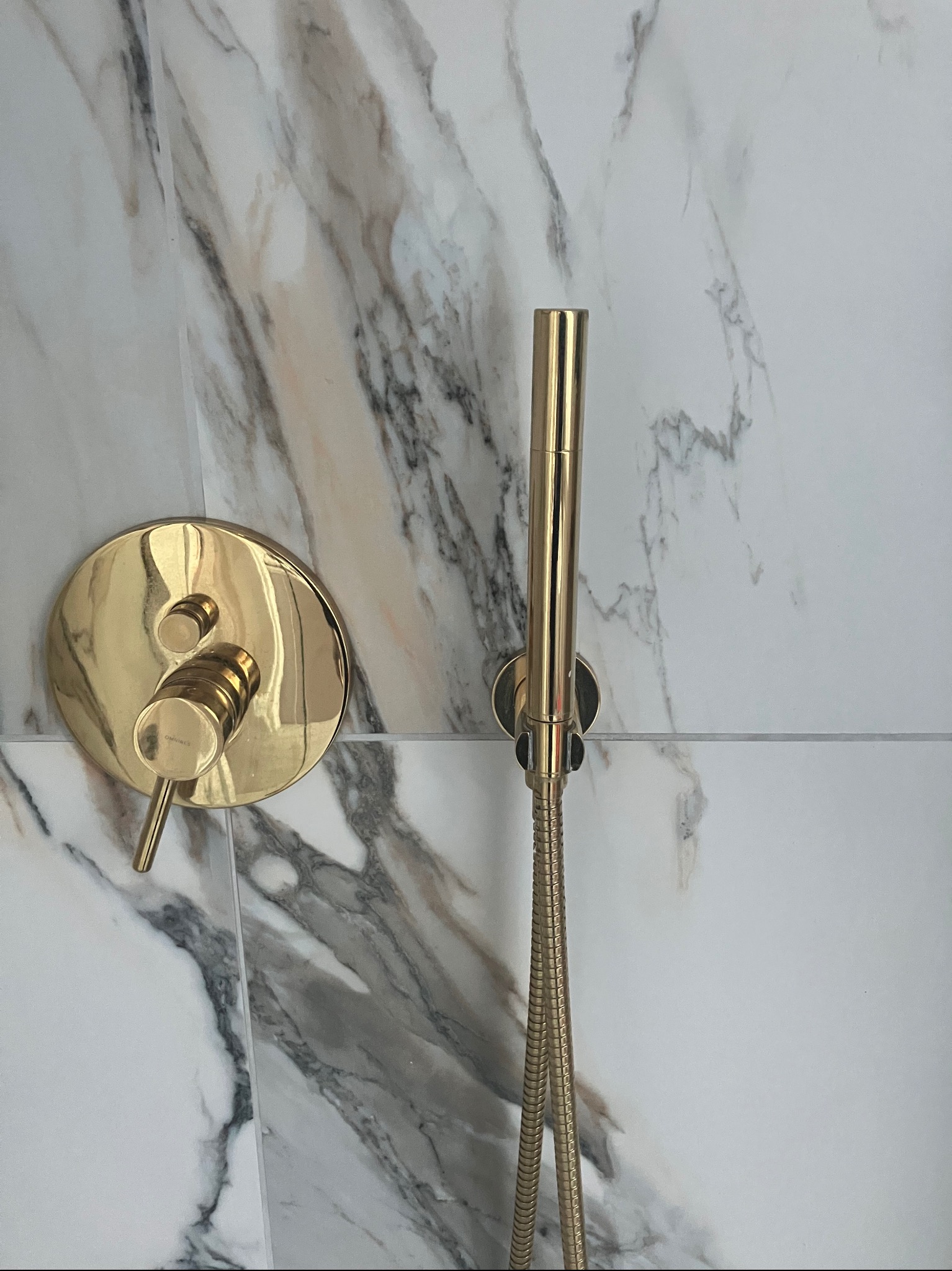 Błyszcząca złota bateria prysznicowa z wężem i okrągłym panelem na ścianie wyłożonej marmurowymi płytkami.