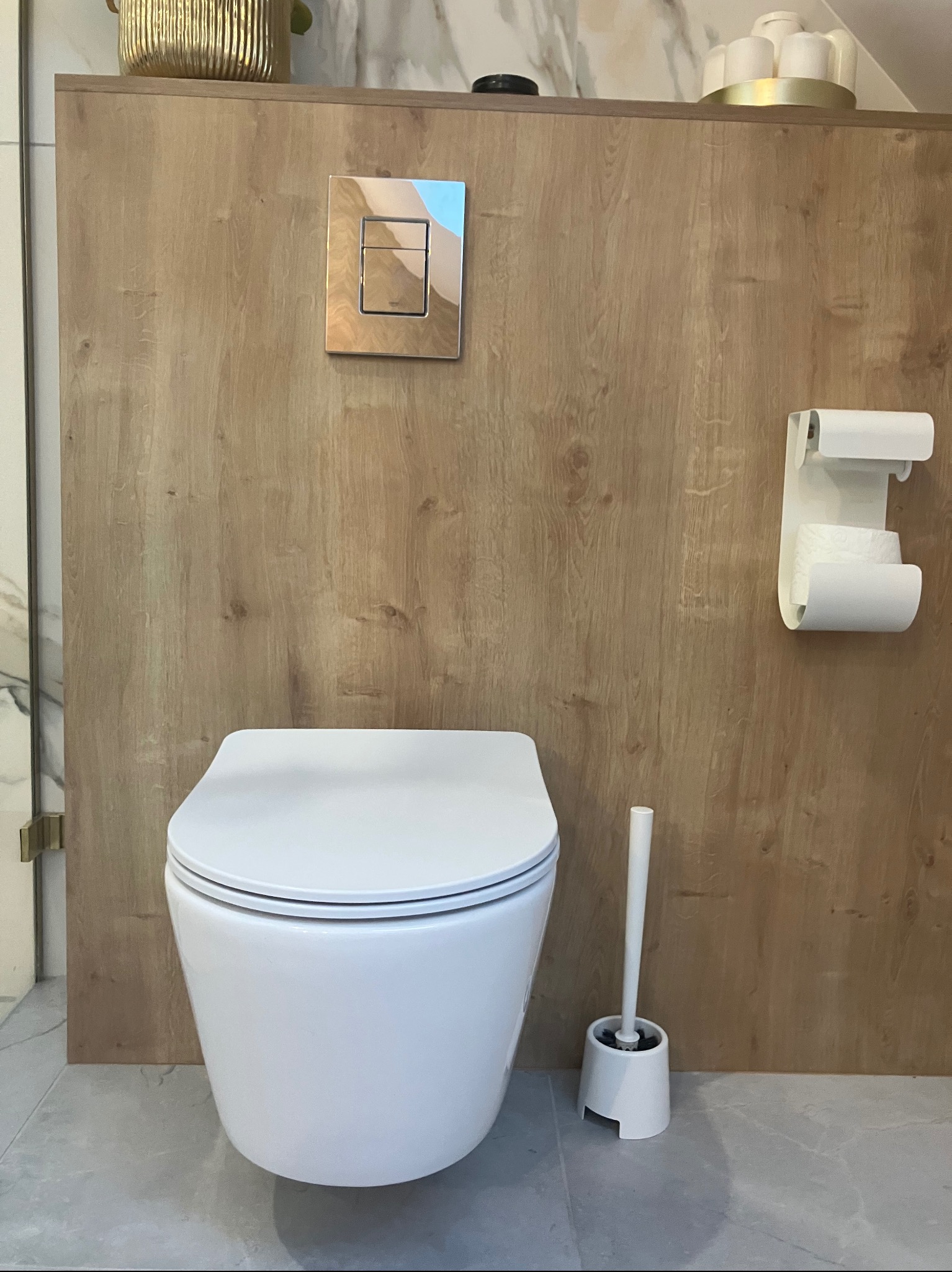 Nowoczesna toaleta podwieszana z drewnianą okładziną ściany, chromowanym przyciskiem spłukiwania i białym uchwytem na papier toaletowy, uzupełniona szczotką do WC w minimalistycznym stylu.
