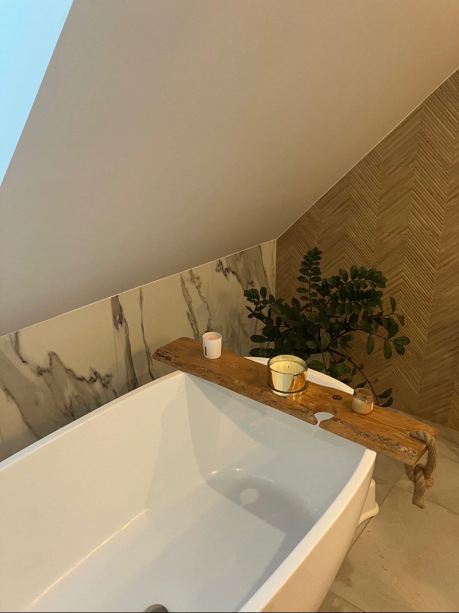Biała wanna wolnostojąca z drewnianą półką z linami, na której stoją świeczki i roślina, na tle marmurowych i drewnianych płytek w łazience na poddaszu.