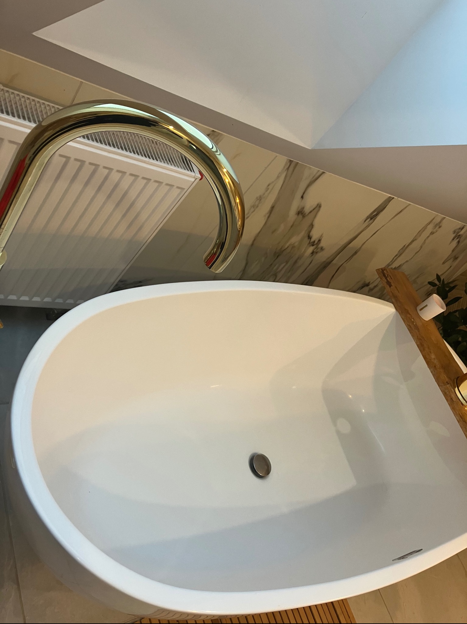 Owalna, biała wanna z złotą baterią, marmurowe płytki na ścianie, drewniana półka z akcesoriami do kąpieli, widok z góry.