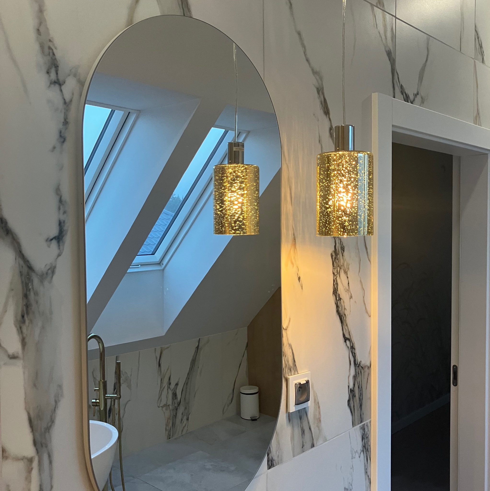 Nowoczesna łazienka z marmurowymi płytkami, owalnym lustrem odbijającym okno dachowe i złotymi lampami wiszącymi