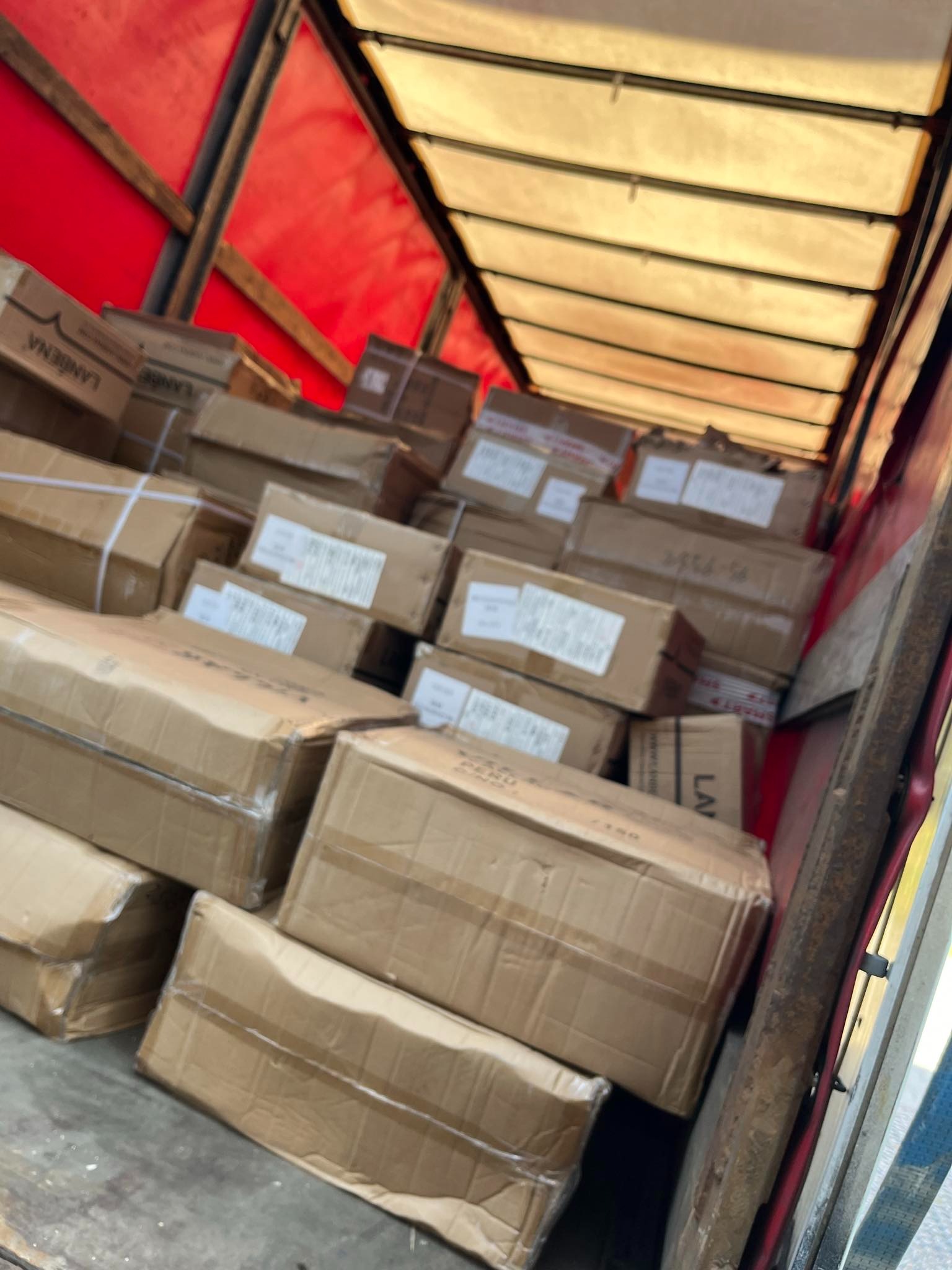 Wnętrze ciężarówki wypełnionej kartonami z taśmą, widoczne etykiety z napisami, czerwona plandeka na dachu