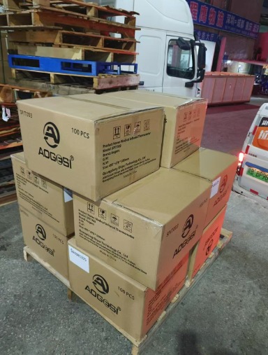 Paleta z kartonami termometrów bezdotykowych AOGESI, gotowa do transportu, na tle ciężarówki i magazynu.