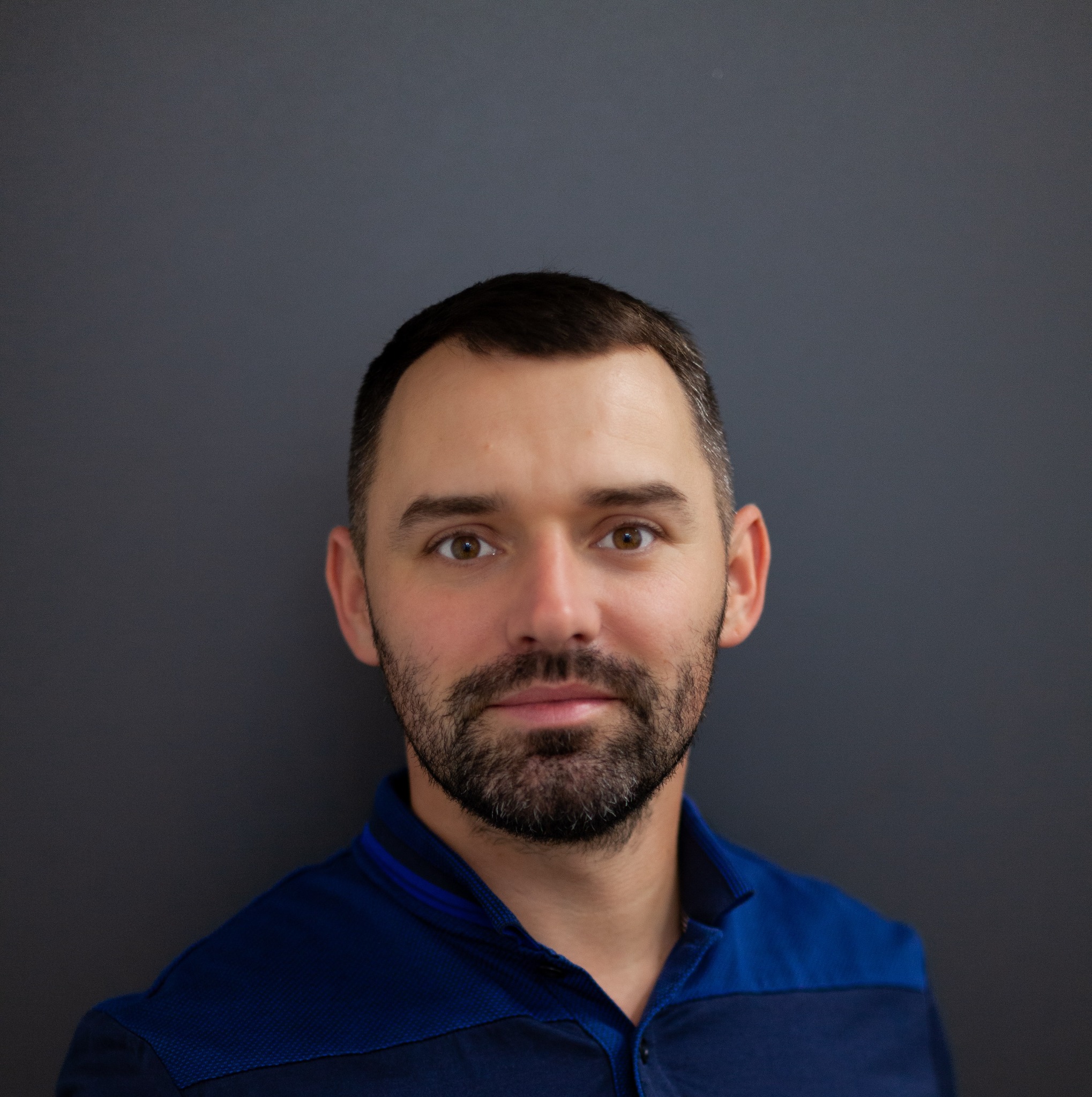 Portret mężczyzny z krótką brodą i brązowymi oczami, ubrany w niebieską koszulę polo, na szarym tle.