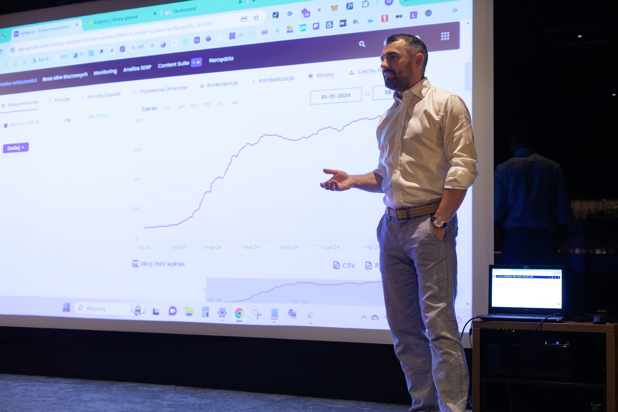 Prelegent w koszuli i spodniach, gestykulujący przed ekranem z wykresem analitycznym strony internetowej, widoczne zakładki przeglądarki z narzędziami SEO i analizą konkurencji, obok laptop...