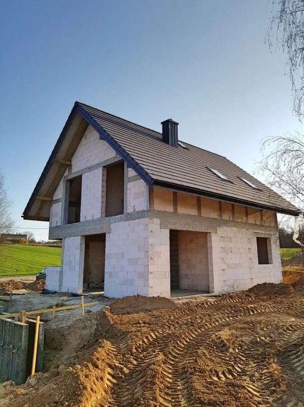 Surowy stan domu jednorodzinnego z białego betonu komórkowego, z zamontowanym dachem z brązowej dachówki ceramicznej i czarnym kominem, widoczne ślady opon ciężkiego sprzętu na ziemi.