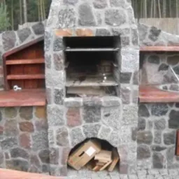 Kamienny grill ogrodowy z paleniskiem, rusztem, drewnianymi półkami i schowkiem na akcesoria, otoczony kostką brukową i zielenią.