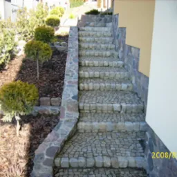 Zewnętrzne schody z kostki granitowej prowadzące do budynku, z kamiennymi obrzeżami i roślinnością po lewej stronie, sfotografowane w słoneczny dzień.