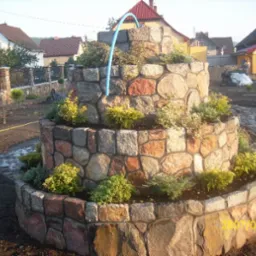 Trzykondygnacyjna fontanna ogrodowa z nieregularnych kamieni polnych, obsadzona zielenią, z niebieską rurką doprowadzającą wodę, na tle ogrodu w trakcie prac wykończeniowych.